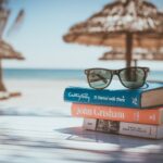 Bild über Bücherstapel am Strand mit Sonnenbrille obendrauf. Im Hintergrund Sonnenschirme