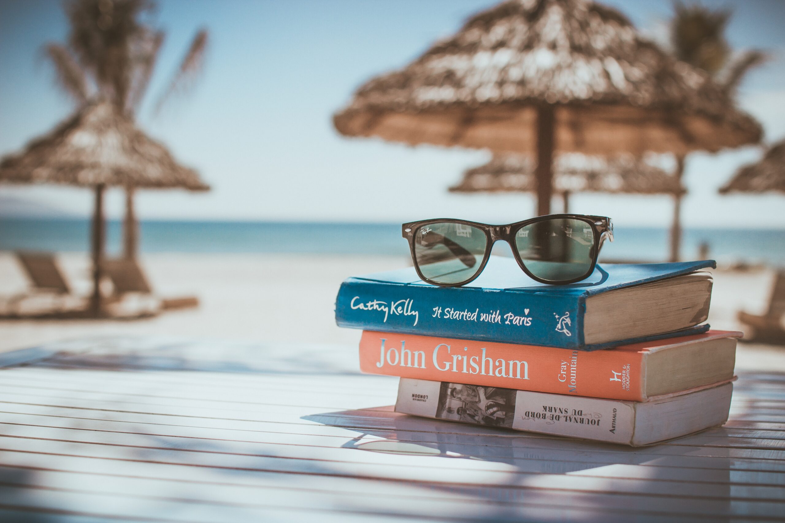 Bild über Bücherstapel am Strand mit Sonnenbrille obendrauf. Im Hintergrund Sonnenschirme